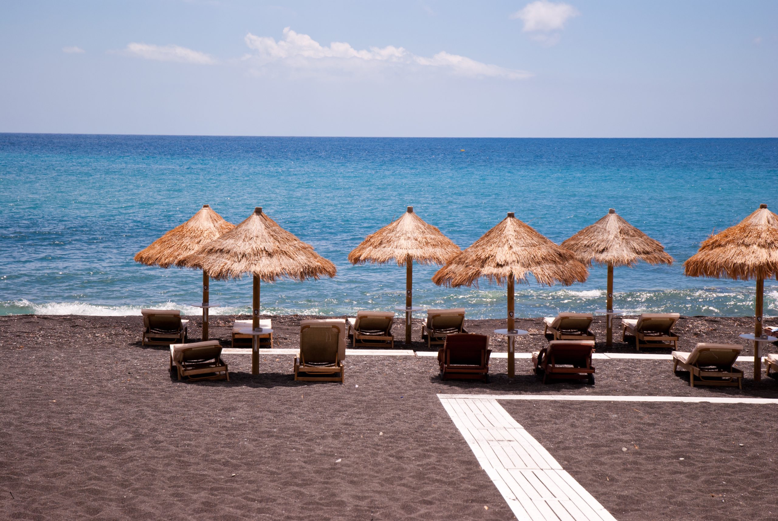 Straw-thatched umbrellas and wooden loungers facing the blue Aegean Sea on Santorini's black sand beach. 