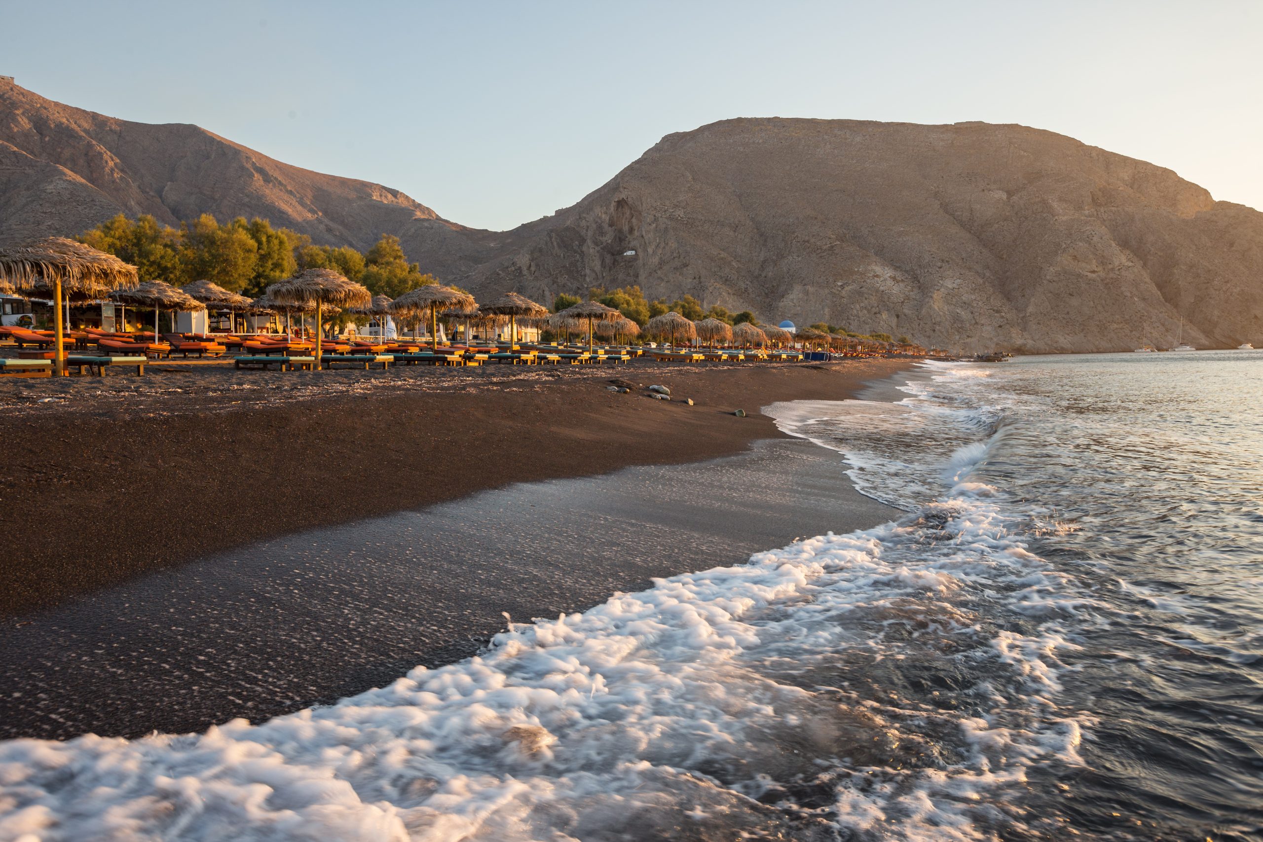 Waves washing onto Perissa's black sand beach at sunrise, lined with colorful sun loungers beneath rocky hills. 