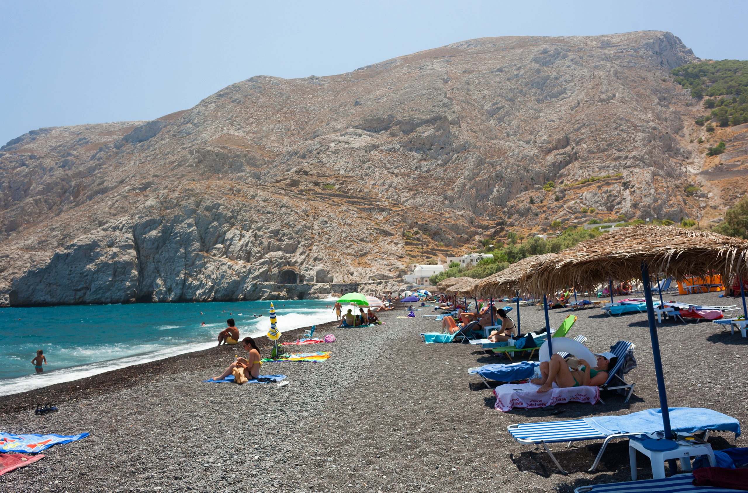 Pebble beach on Santorini with sun loungers and swimmers beneath rugged grey cliffs.