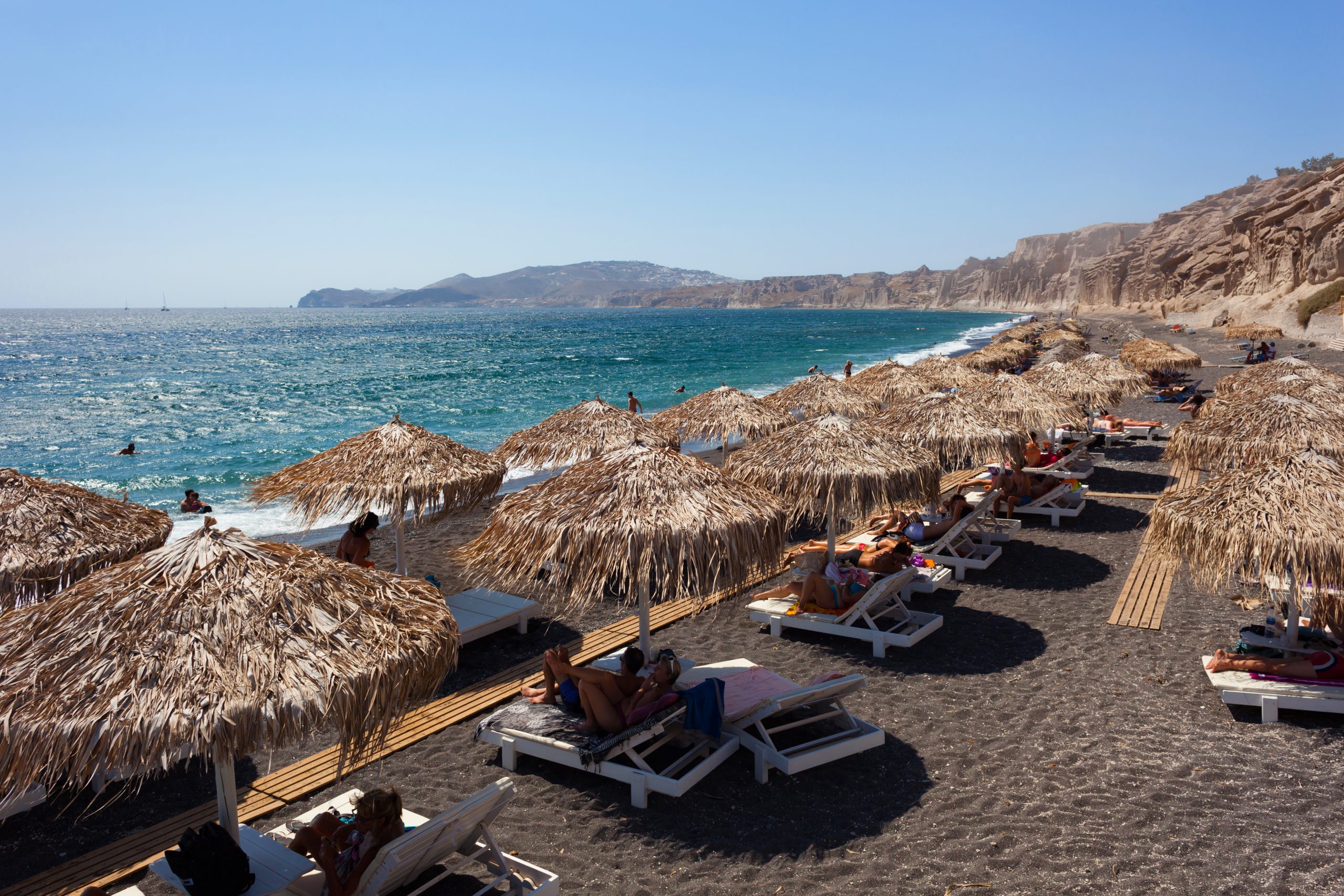 Straw-thatched beach umbrellas lining Santorini's black volcanic sand shore, with sunbathers overlooking the turquoise Aegean.