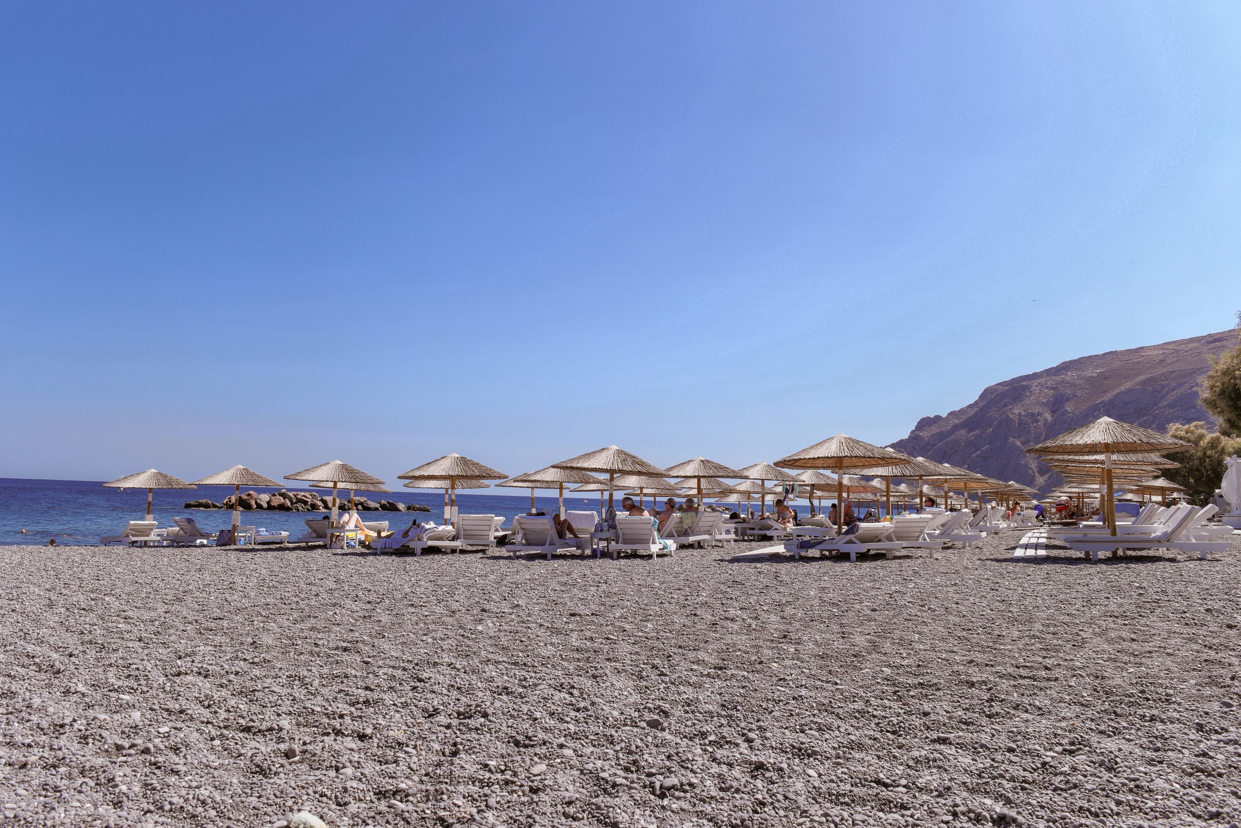 Straw beach umbrellas and sun loungers on Perissa's grey pebble beach, with cliffs in the distance. 