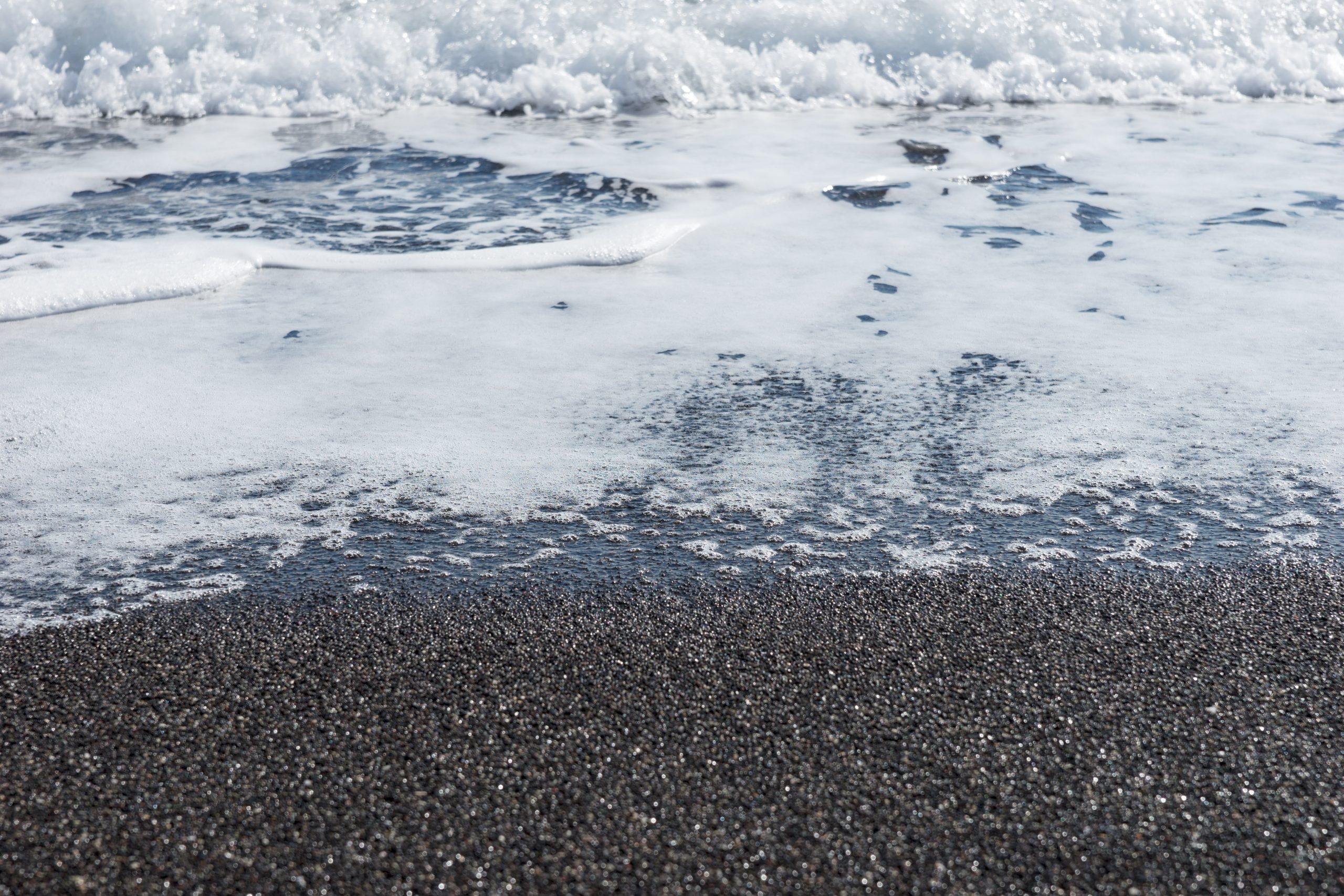 Close-up of foamy waves washing over Santorini's black volcanic sand beach.