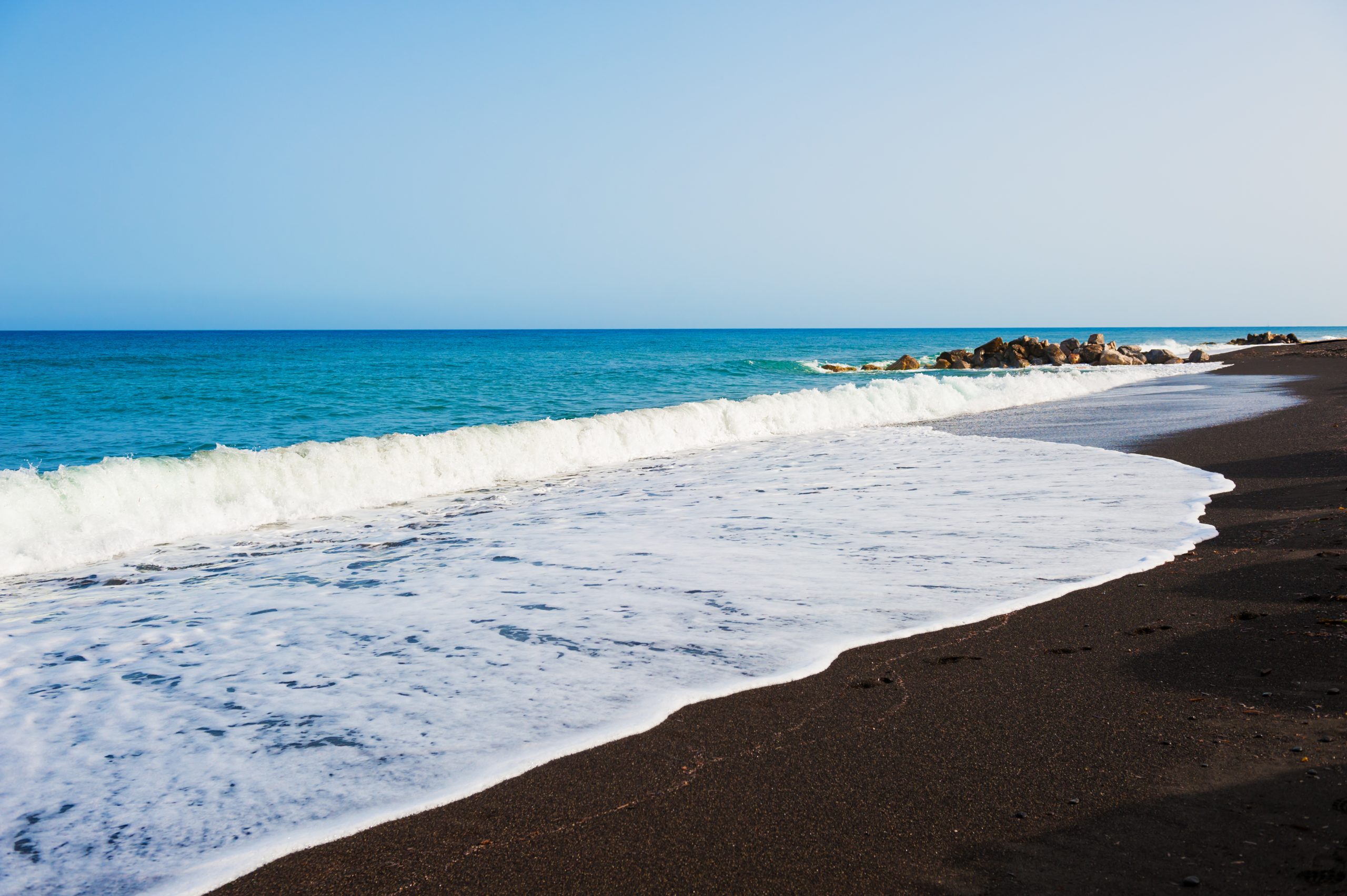 Turquoise waves breaking onto Santorini's black sand beach, with rocks lining the shore.