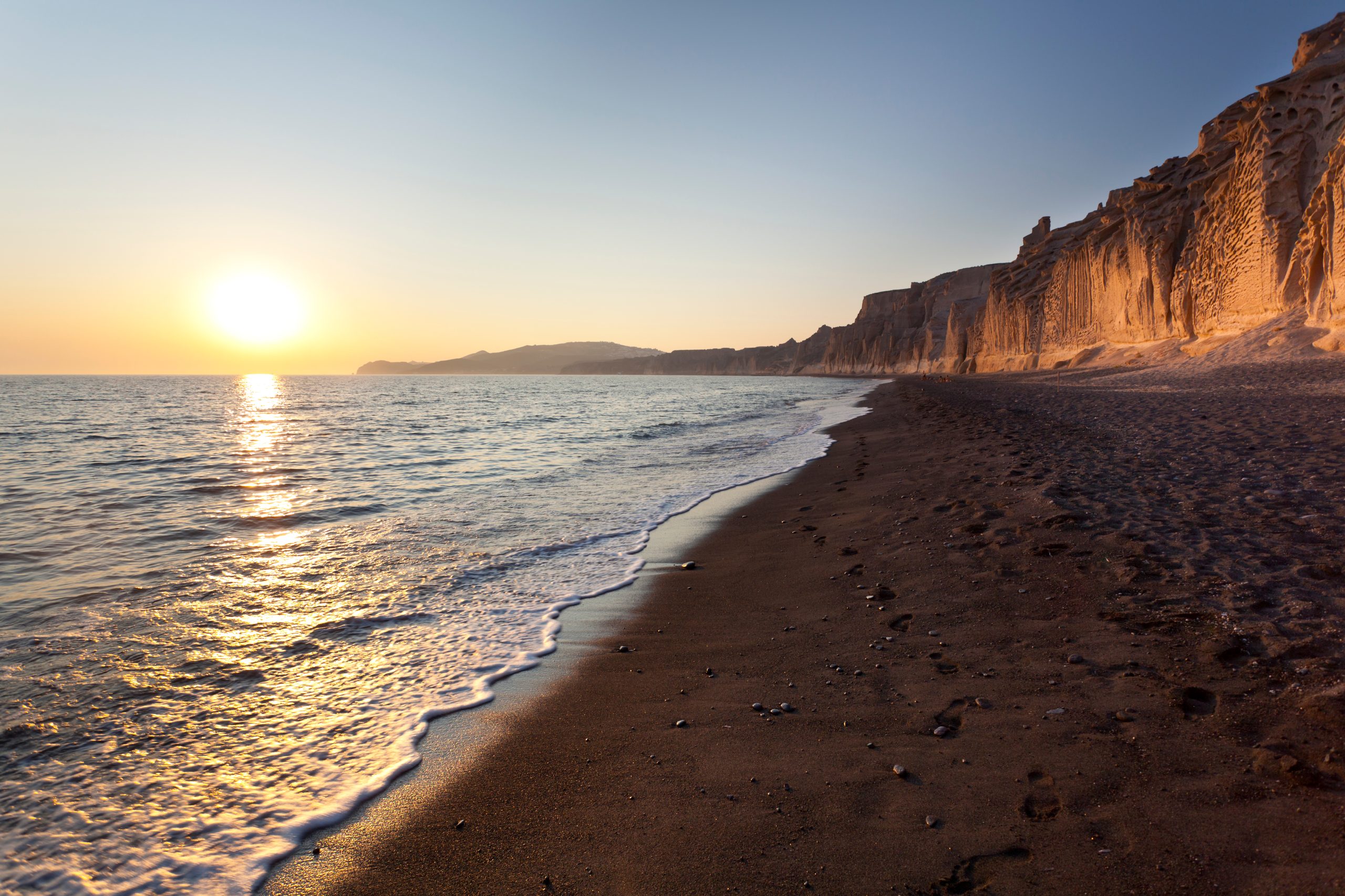 Sunset over Vlychada Beach, Santorini, with dramatic sculpted cliffs and dark volcanic sand along the shore.