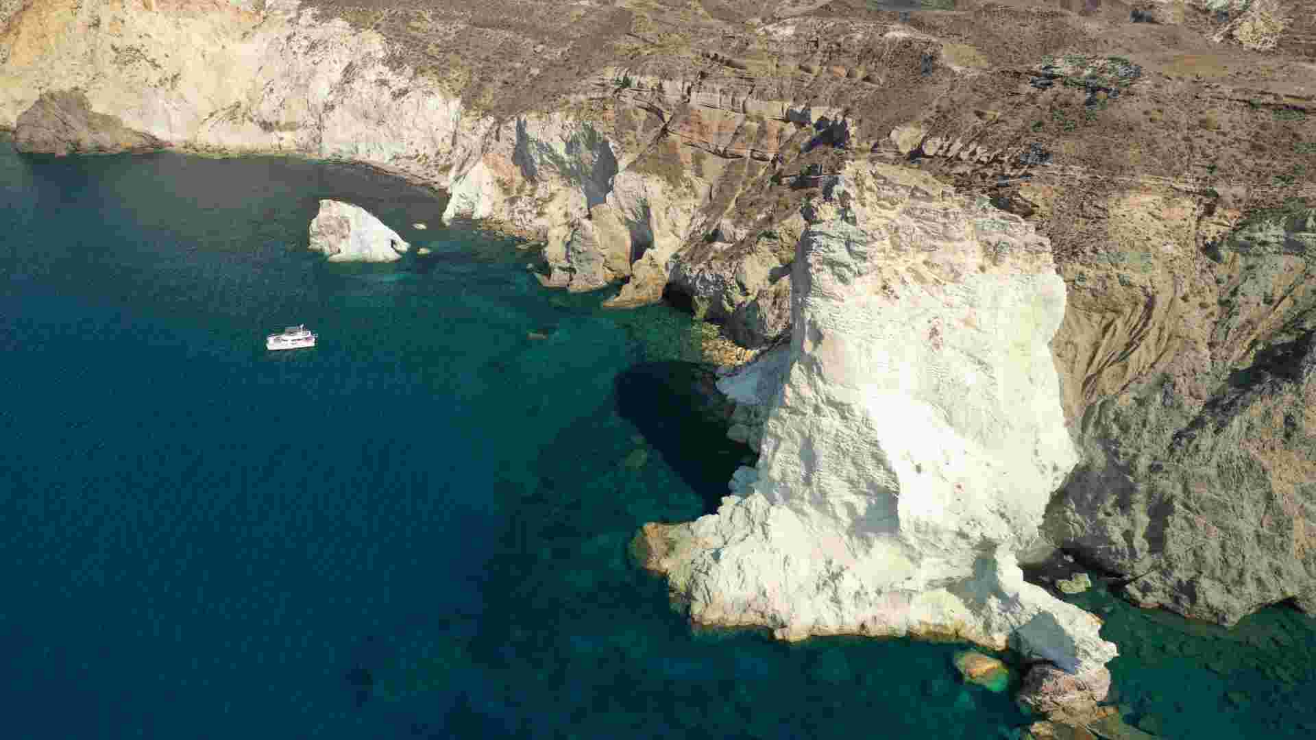 Aerial view of white volcanic rock formations and sea caves along Santorini's coastline, with a boat anchored offshore. Aerial,Drone,Photo,Of,Iconic,Famous,White,Beach,With,Deep