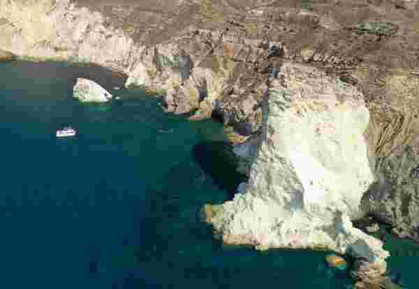 Aerial view of white volcanic rock formations and sea caves along Santorini's coastline, with a boat anchored offshore. Aerial,Drone,Photo,Of,Iconic,Famous,White,Beach,With,Deep