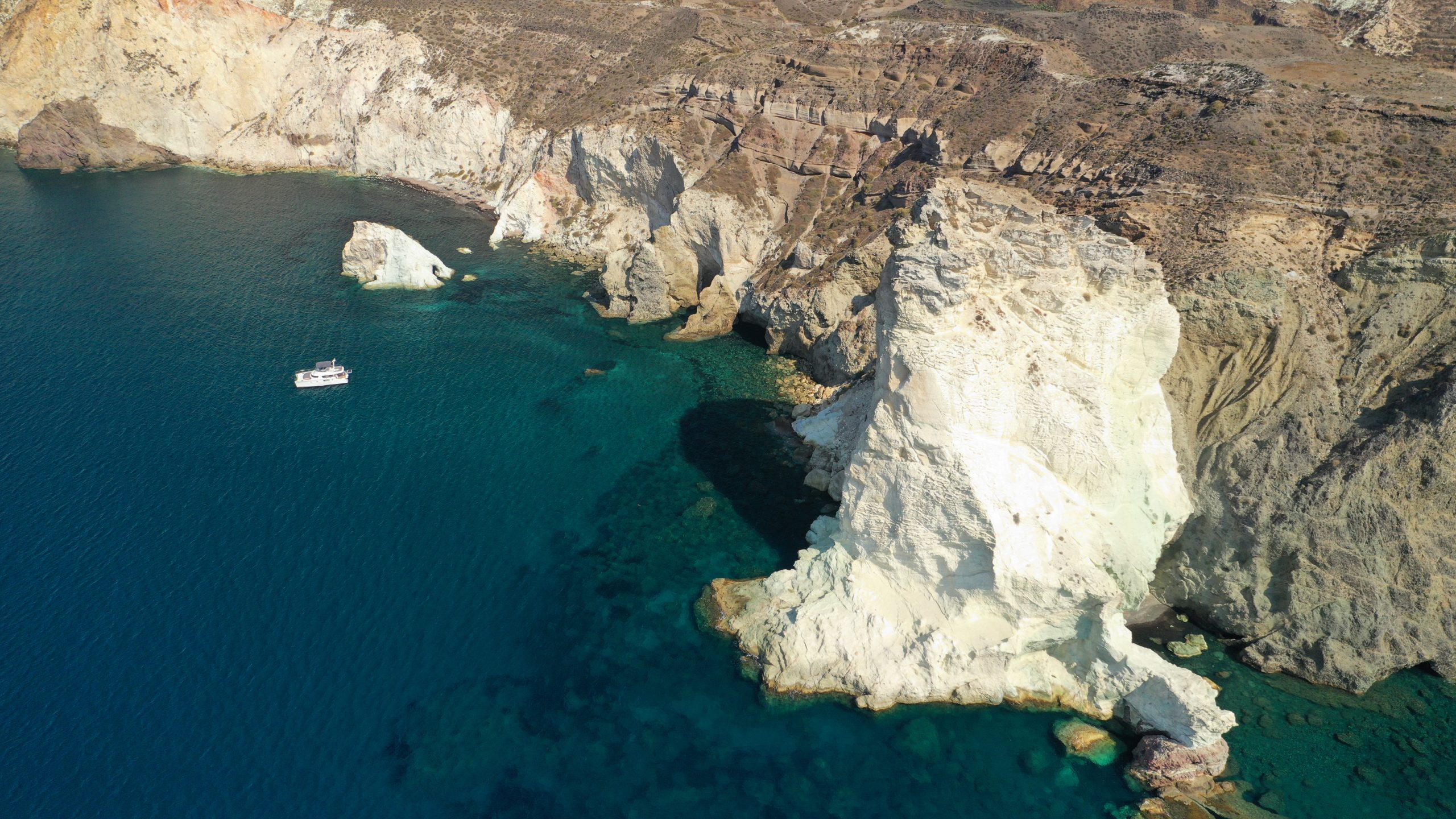 Aerial view of white volcanic rock formations and sea caves along Santorini's coastline, with a boat anchored offshore.