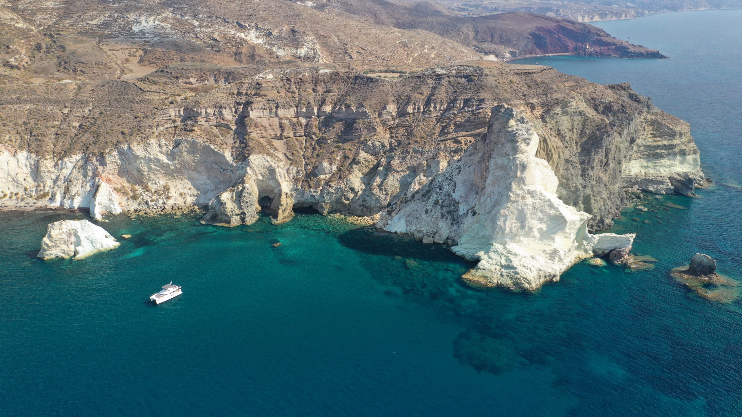Aerial view of Santorini's white rocky coastline with sea caves and a boat floating in clear turquoise water.