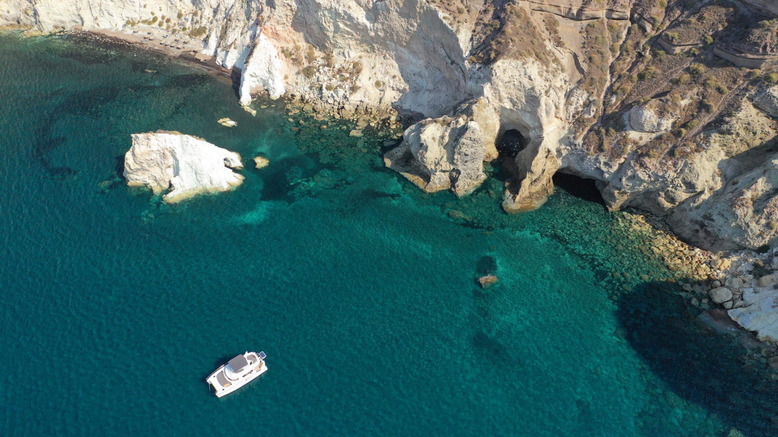 Aerial view of a boat anchored near white cliffs and a hidden sea cave along Santorini's coast.
