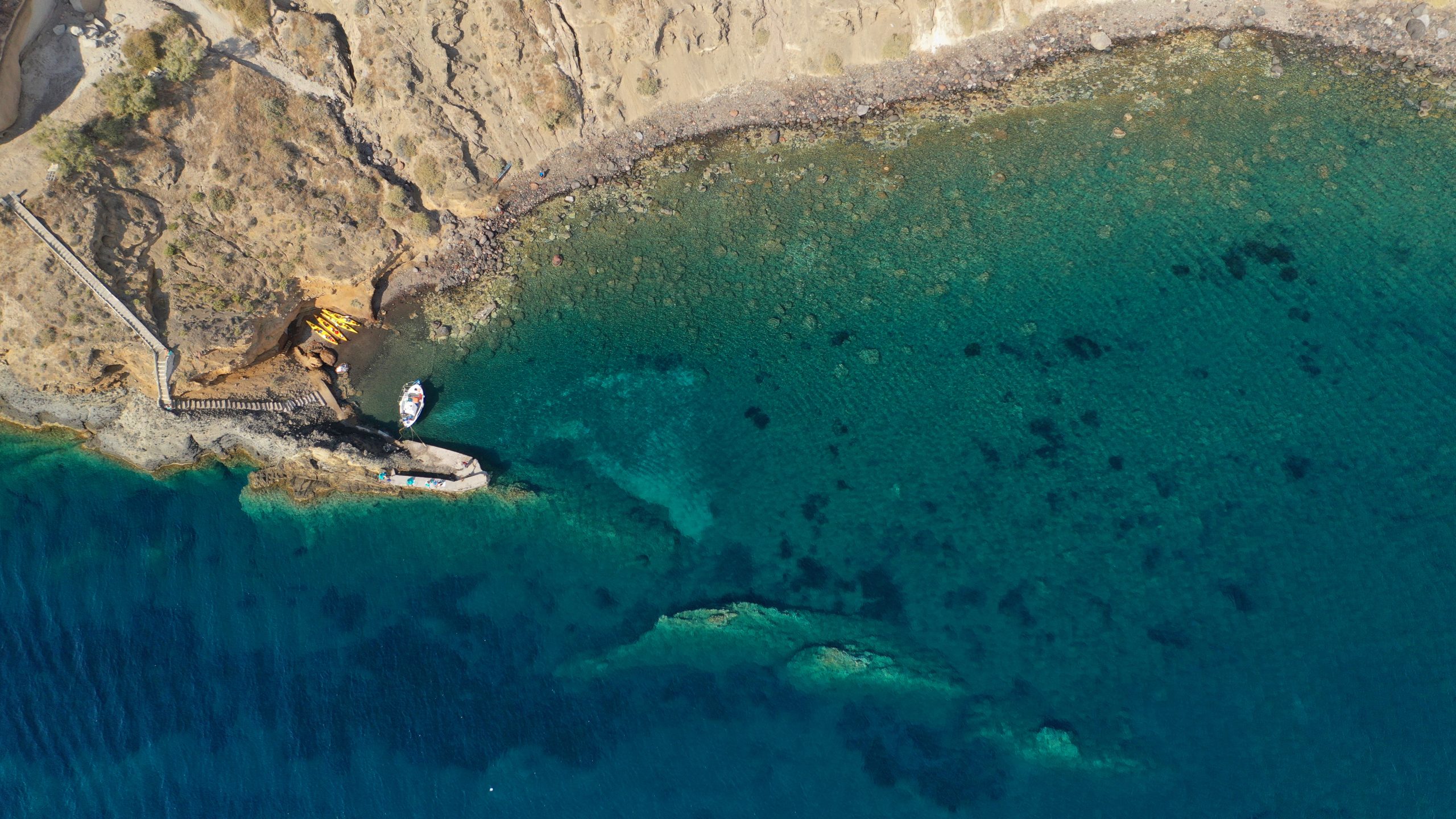 Aerial view of a small Santorini cove with kayaks and a moored boat beside turquoise water.