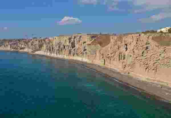 Aerial view of Santorini's white pumice cliffs rising above a dark sand beach and turquoise sea. Aerial,Drone,Photo,Of,Amazing,Shape,Giant,Volcanic,Rock,Formations