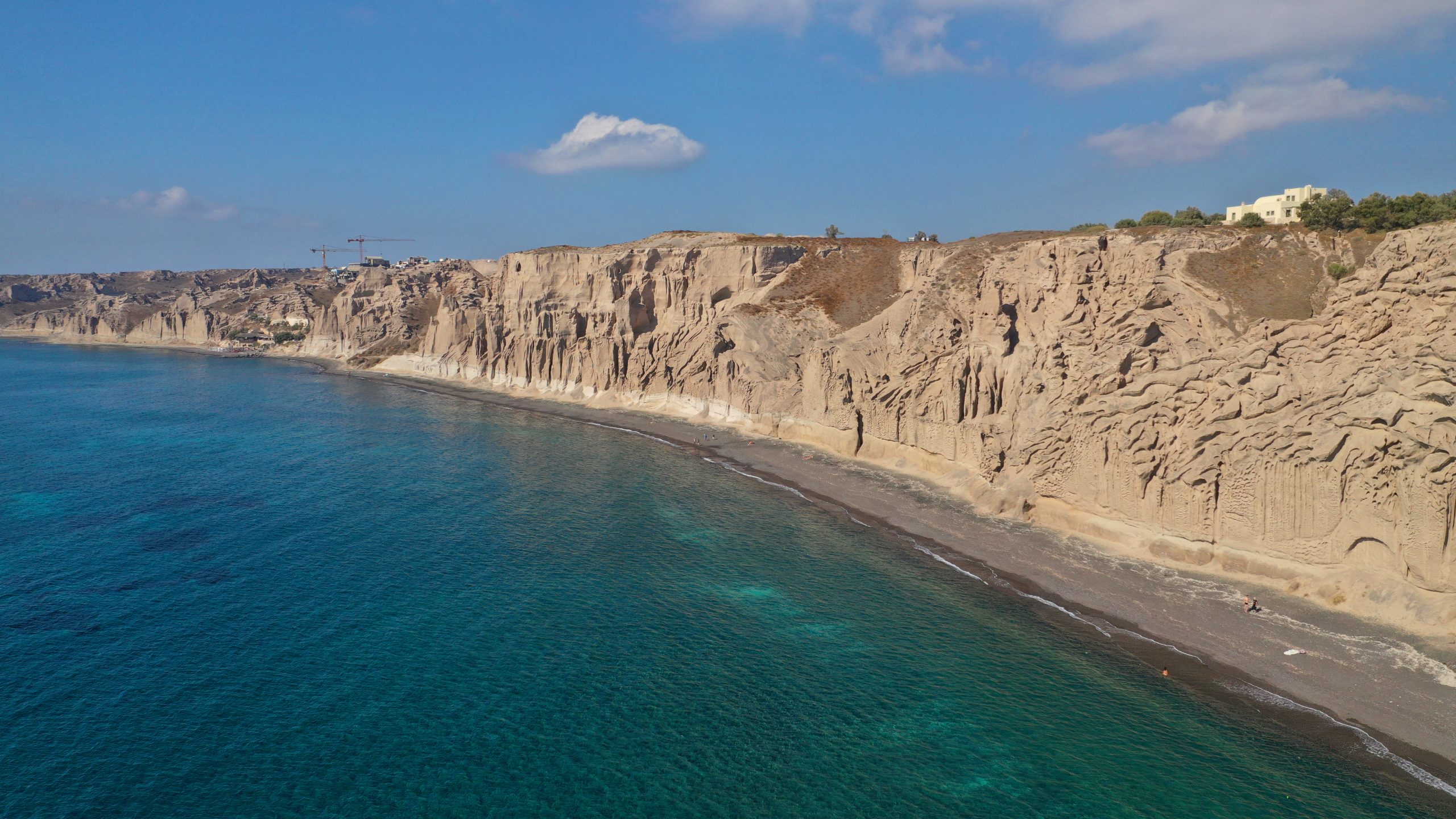 Aerial view of Santorini's white pumice cliffs rising above a dark sand beach and turquoise sea. 