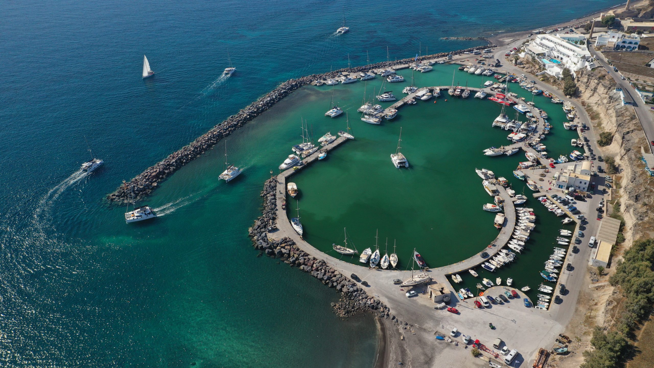 Aerial view of a circular marina on Santorini, with yachts and sailboats moored in turquoise water. 