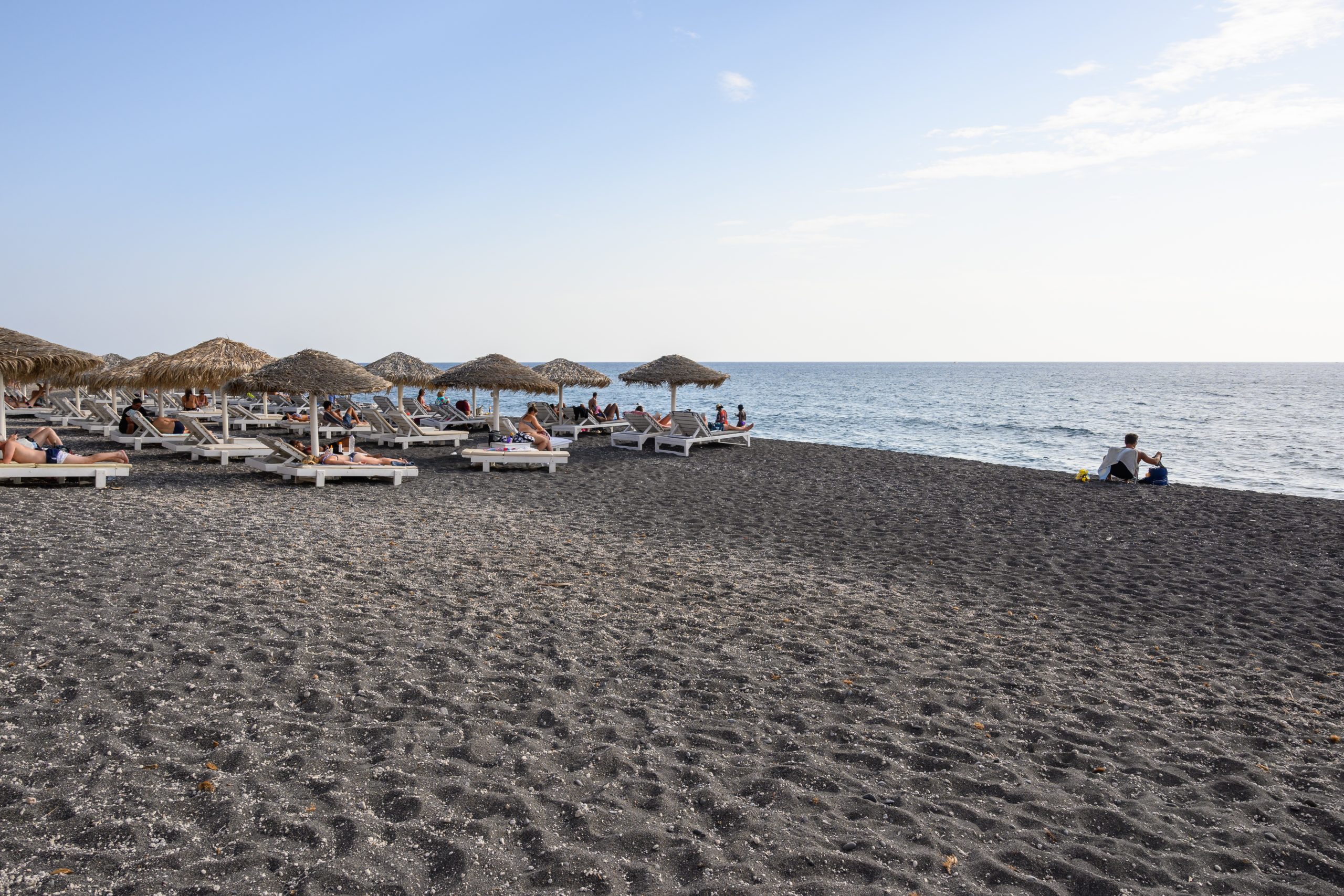 Straw-thatched umbrellas and sun loungers lining Santorini's black sand beach, with the calm sea beyond. 
