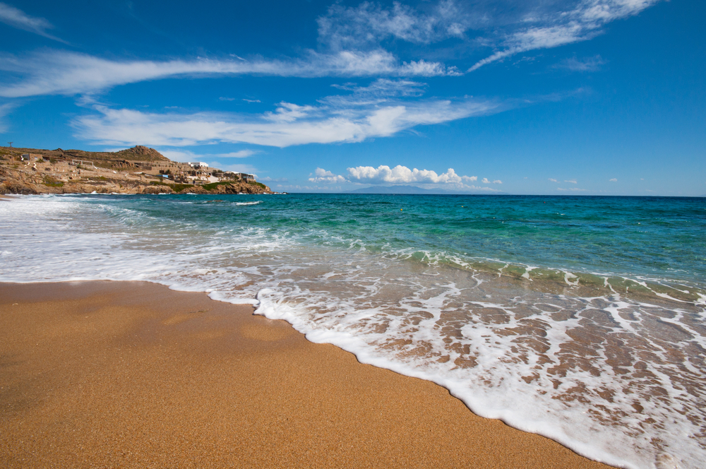 Frothy waves wash over a golden sand beach in Mykonos, Greece, with a rocky headland and whitewashed buildings in the distance under a partly cloudy sky.