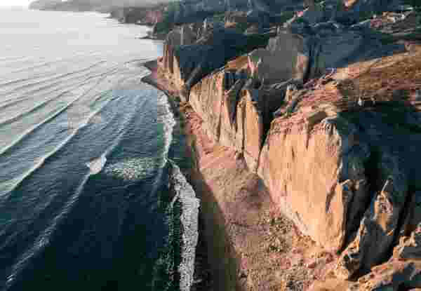 Aerial view of Santorini's sculpted volcanic cliffs meeting a narrow black sand beach, waves rolling in along the coastline. A,Beautiful,View,Of,The,Eros,Beach,,Santorini,,Greece