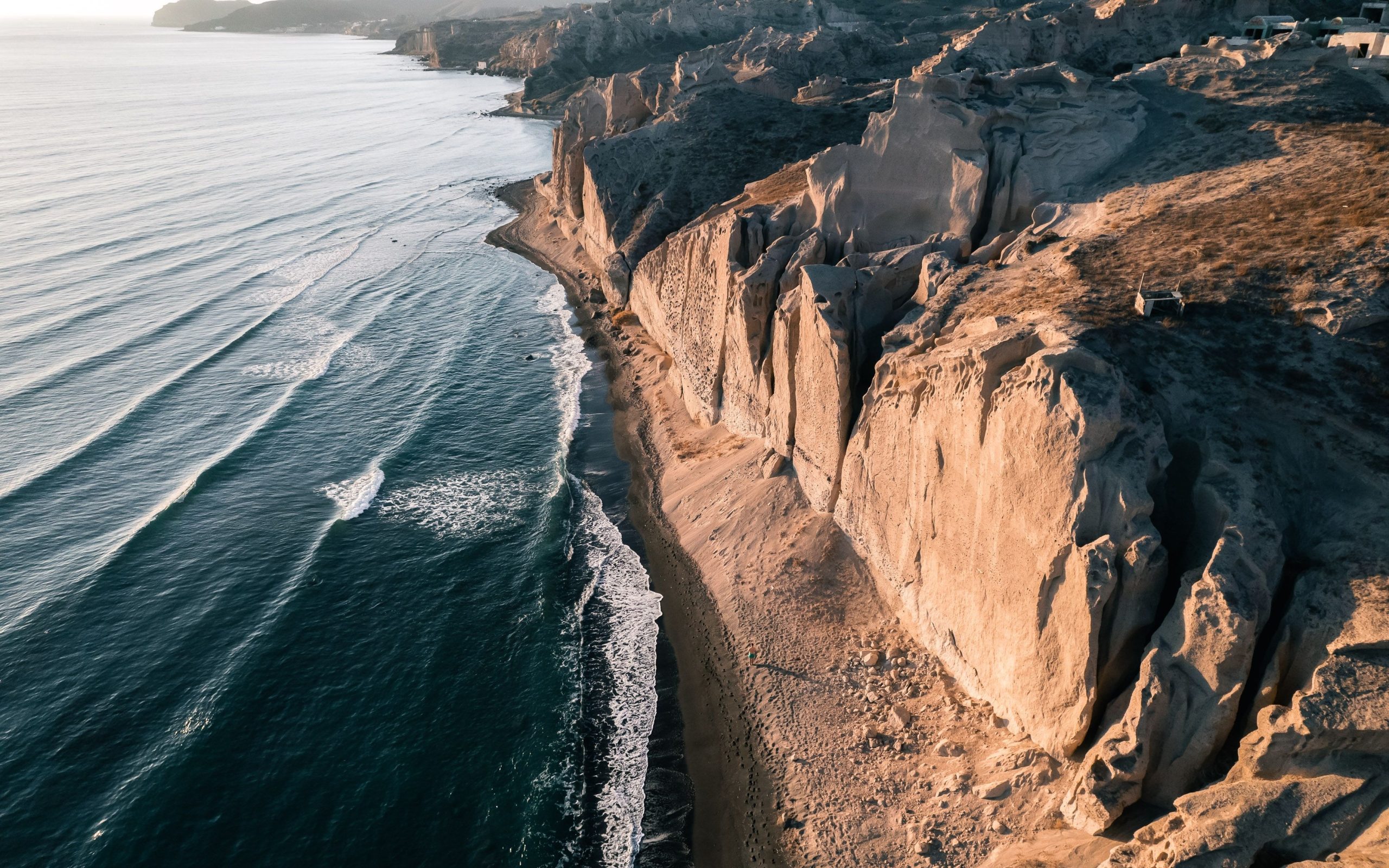 Aerial view of Santorini's sculpted volcanic cliffs meeting a narrow black sand beach, waves rolling in along the coastline.