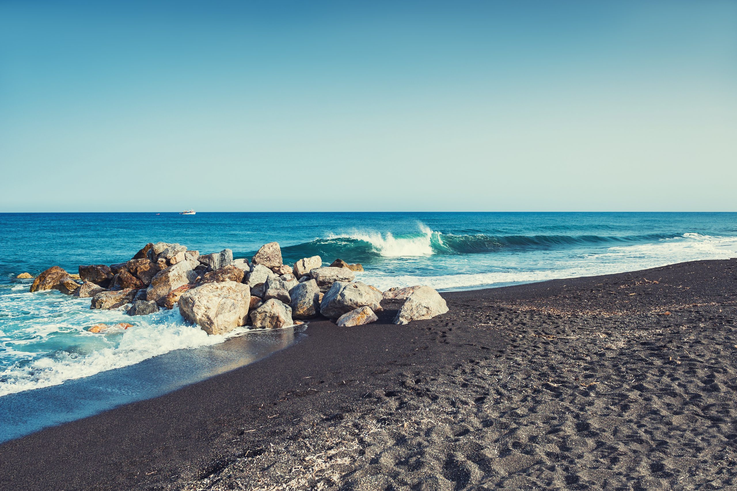 Waves crashing over rocks on a black sand beach, with a fishing boat visible on the horizon.
