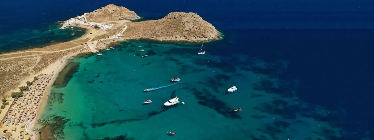 Aerial view of Agia Anna, Mykonos, with rocky coastline and yachts anchored in turquoise water beside golden sand. Agia Anna Beach Mykonos 1