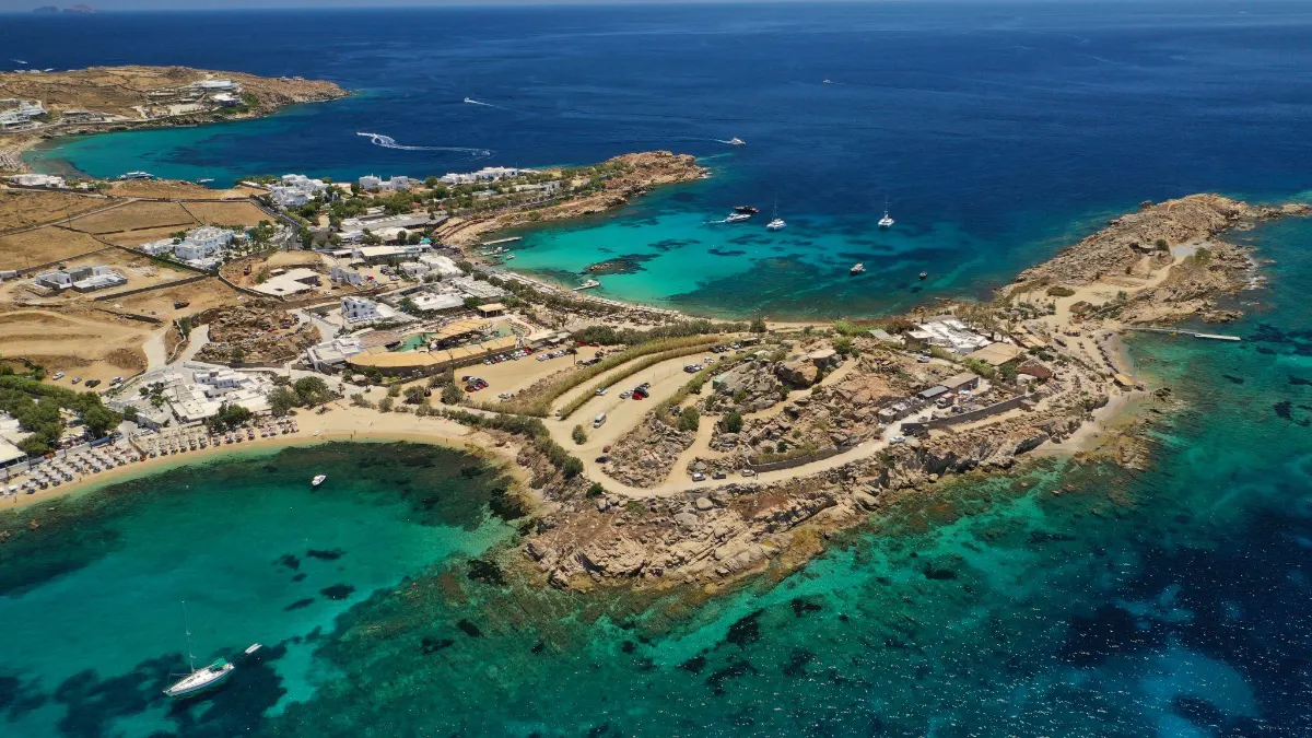 Aerial view of Agia Anna Mykonos's rocky coastline, with turquoise coves, beach clubs, and yachts anchored between sandy inlets and sunbaked headlands.
