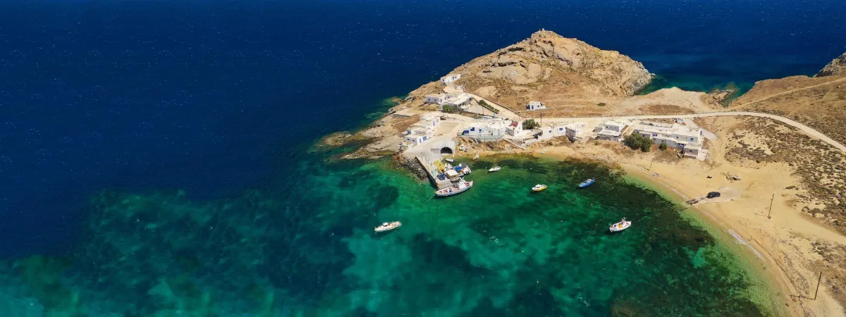 Aerial view of Agia Anna, Mykonos, with yachts and sailboats anchored in a turquoise bay, backed by whitewashed hillside villages.