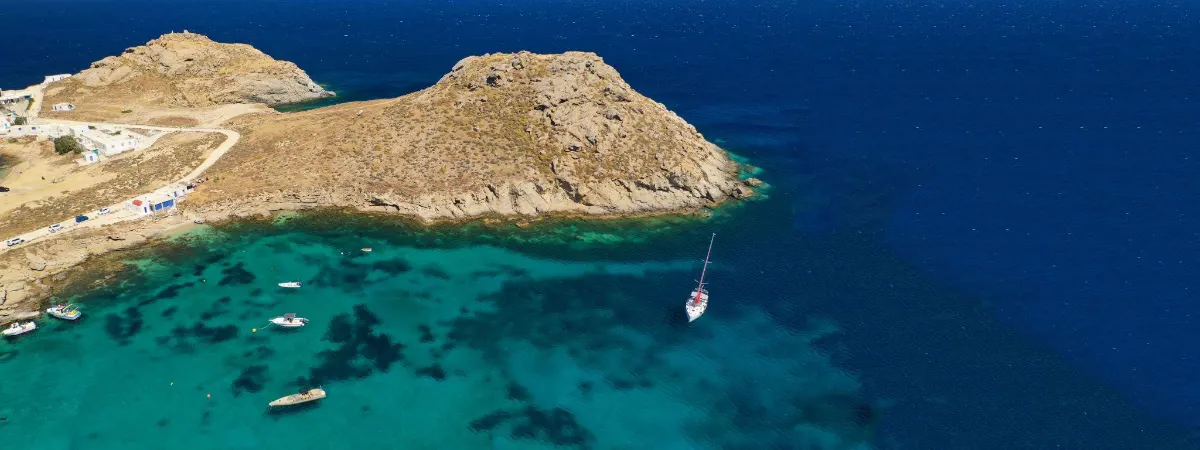Aerial view of Agia Anna bay in Mykonos, Greece, with yachts moored in turquoise water beside a sandy beach lined with sunbeds and beach clubs.