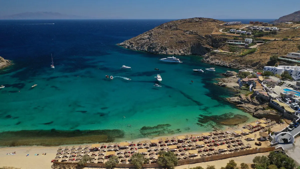 Aerial view of Agia Anna beach in Mykonos, Greece, with yachts anchored in a turquoise bay and whitewashed villages spread across the surrounding hills.