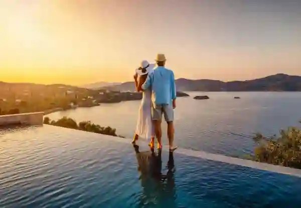 A couple in sun hats stand at the edge of an infinity pool overlooking a Greek coastal bay, with green hills and scattered white villas glowing in the sunset. Greece Honeymoon 1