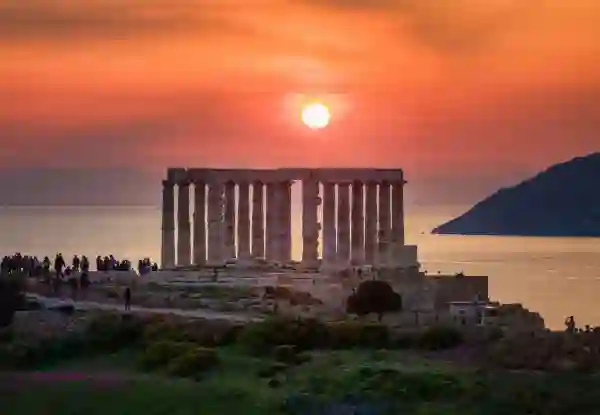The Temple of Poseidon at Cape Sounion, Greece, silhouetted against a fiery orange sunset over the Aegean Sea, with visitors gathered along its ancient marble columns. Greece In Spring 1