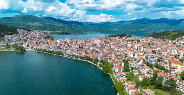 Aerial view of Kastoria, Greece, with red-roofed houses lining the shore of Lake Orestiada, framed by green mountains under a partly cloudy sky. Kastoria 1
