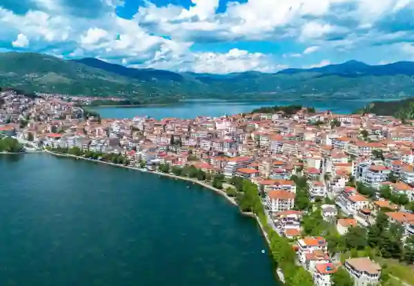 Aerial view of Kastoria, Greece, with red-roofed houses lining the shore of Lake Orestiada, framed by green mountains under a partly cloudy sky. Kastoria 1