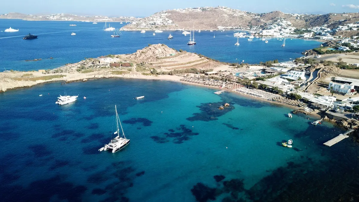 Aerial view of Paraga Beach, Mykonos, with yachts and sailboats anchored in a turquoise bay, backed by whitewashed hillside villages.