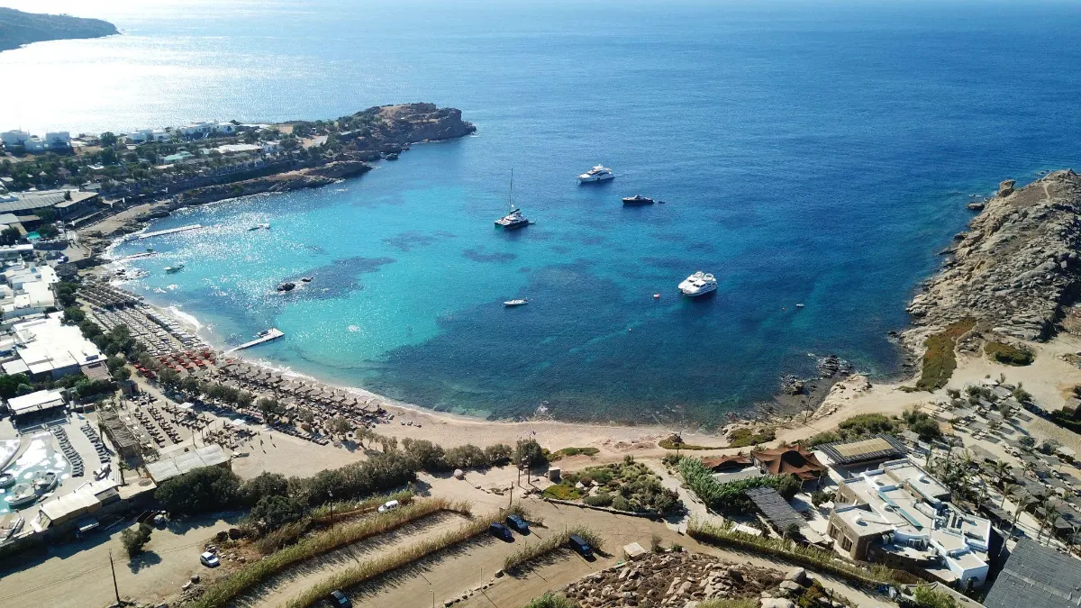 Aerial view of a rocky bay in Mykonos, Greece, with yachts moored in turquoise water beside a sandy beach lined with sunbeds and beach clubs.