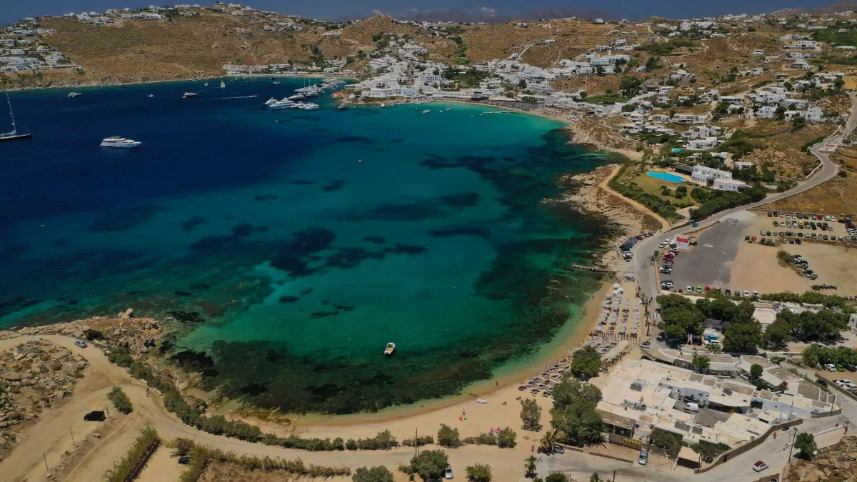 Aerial view of Platis Gialos beach in Mykonos, Greece, with yachts anchored in a turquoise bay and whitewashed villages spread across the surrounding hills.