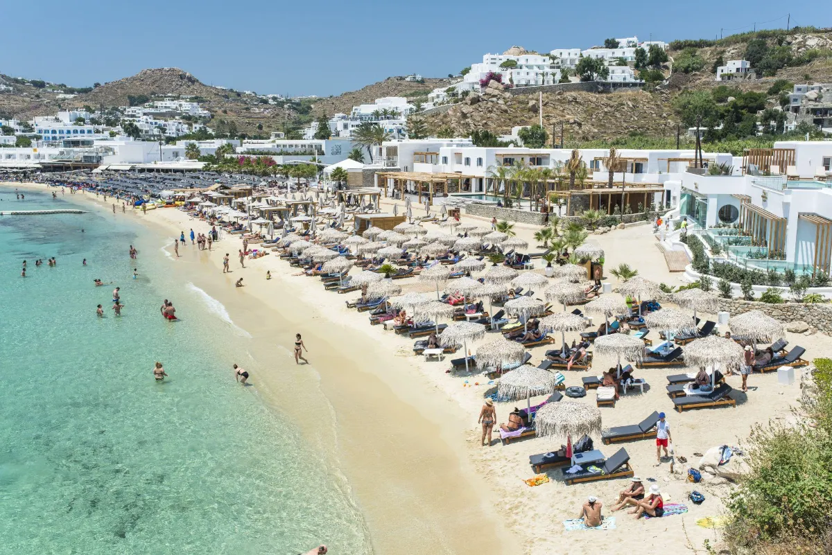 Sunbathers and straw umbrellas on a busy Mykonos beach by clear water