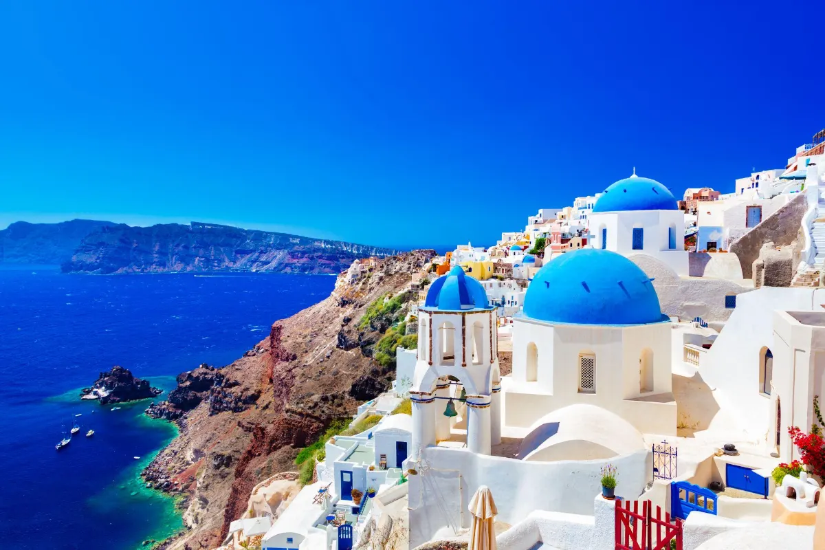Blue-domed churches crown the whitewashed clifftop village of Oia, Santorini, overlooking the deep blue caldera and volcanic coastline. Santorini With Kids 1