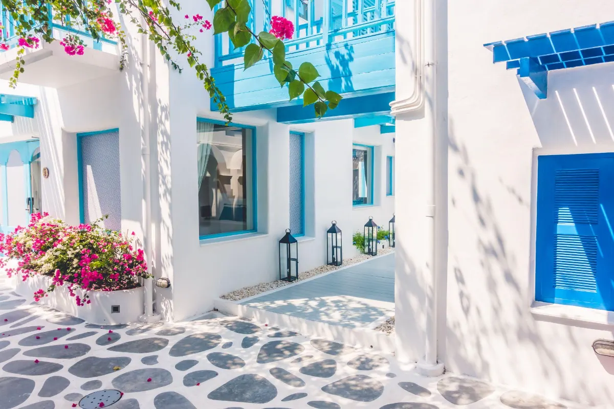 A sunlit Mykonos courtyard with whitewashed walls and bright blue trim, framed by cascading pink bougainvillea and pebble-patterned stone flooring.