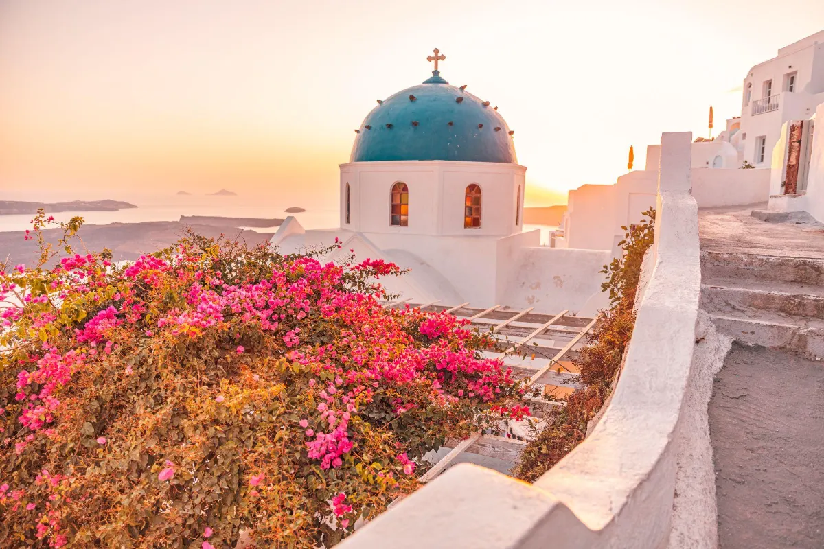 A blue-domed church with stained glass windows overlooks the Santorini caldera at sunset, surrounded by vivid pink bougainvillea.
