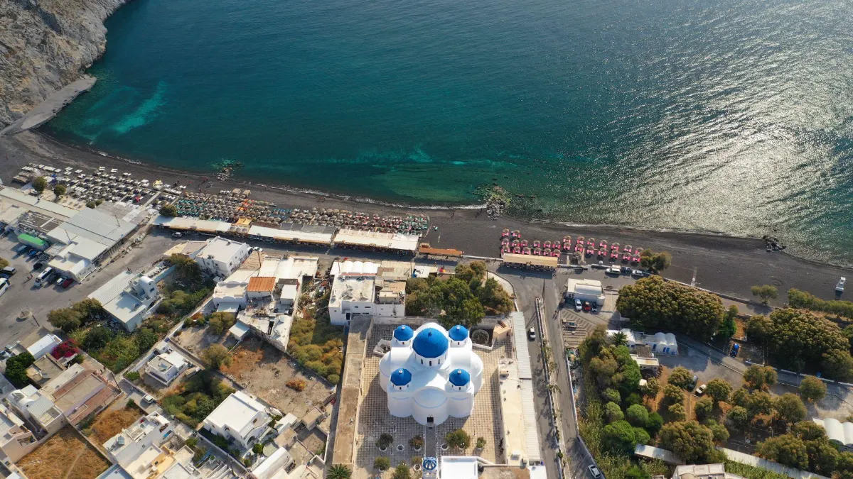 Aerial view of a domed blue and white church in Perissa, Santorini, beside a black sand beach lined with colorful umbrellas along the sea.