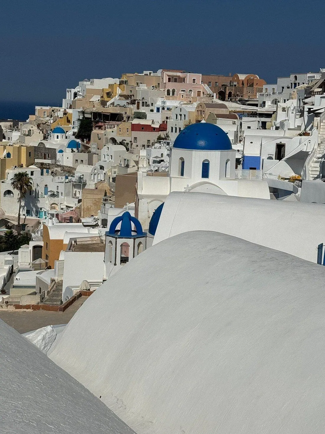 Whitewashed houses and blue-domed churches cascade down the hillside of Oia, Santorini, under a clear blue sky.
