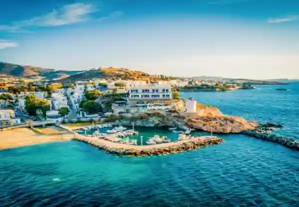 Aerial view of Naoussa harbor in Paros, with a stone windmill and whitewashed hotel overlooking a turquoise fishing port. Rocky breakwaters and colorful wooden boats sit beside a golden sand beach under a clear sky. Things To Do In Paros 1