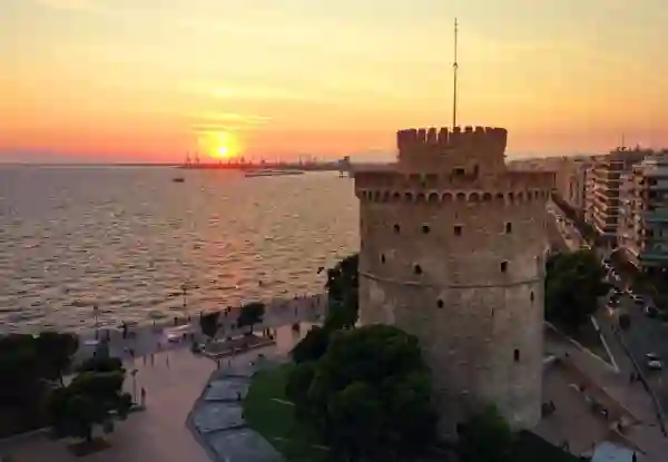 Thessaloniki's White Tower glowing at sunset over the waterfront promenade, with the sun setting over the Thermaic Gulf. Things To Do In Thessaloniki 1