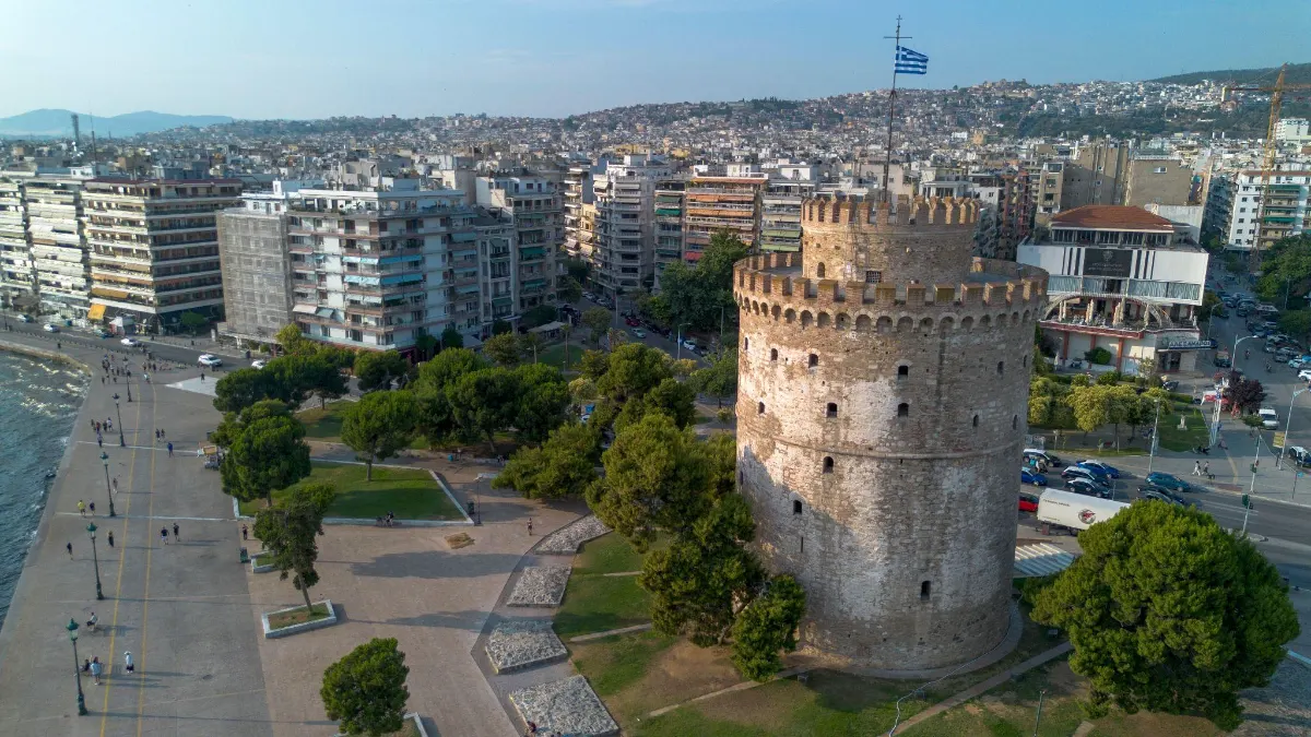 Aerial view of Thessaloniki's White Tower rising above the seaside park, with the sprawling city and hillside neighborhoods behind it.