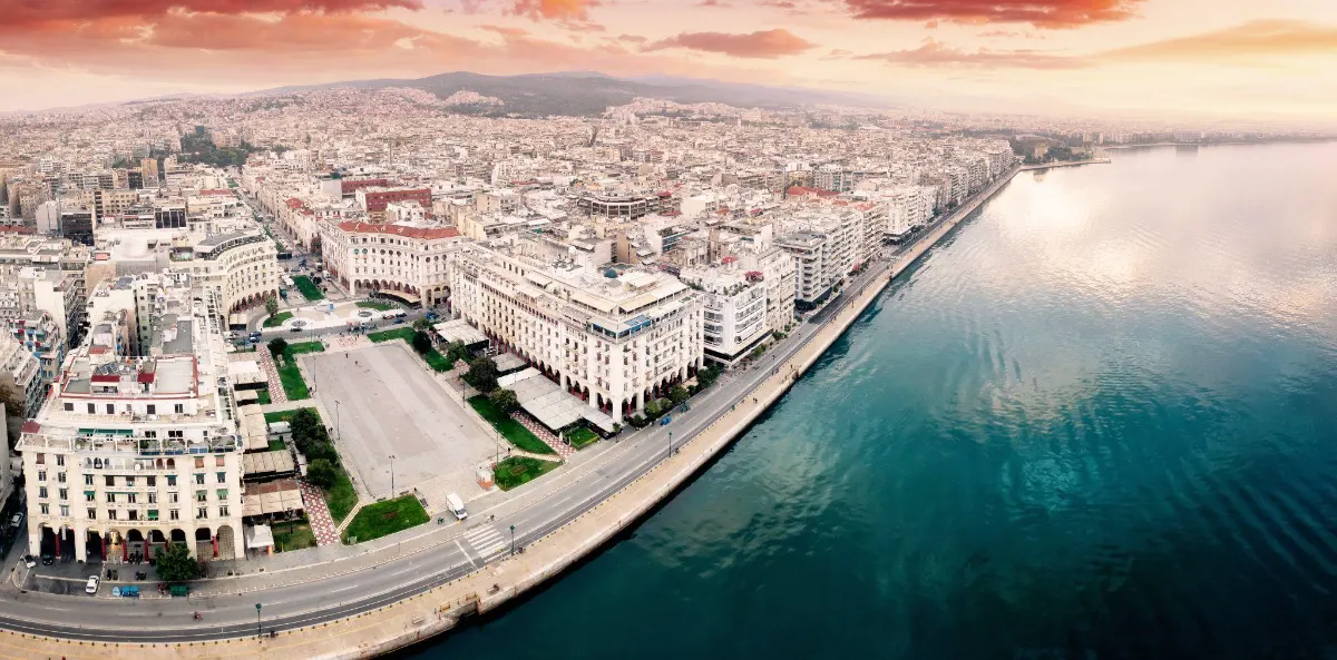 Aerial panorama of Thessaloniki's Aristotelous Square and waterfront promenade curving along the Thermaic Gulf at sunset.