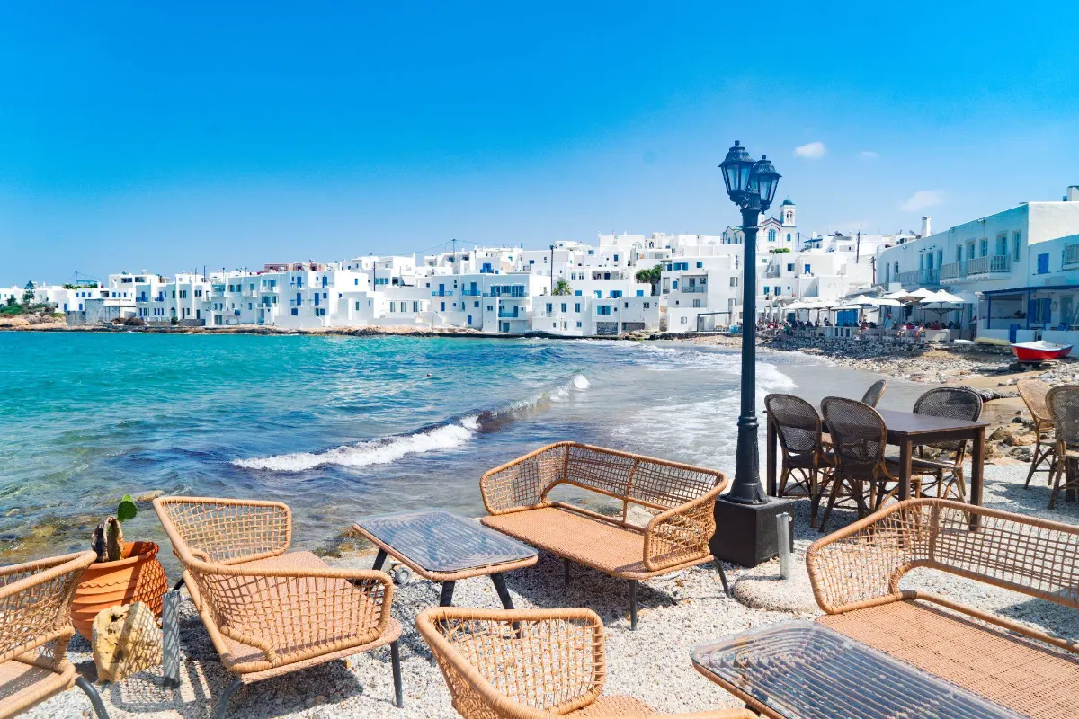 Waterfront cafe seating on the pebbled shore of Naoussa, Paros, overlooking whitewashed houses across the turquoise bay.