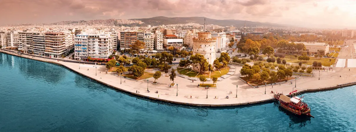 Aerial view of Thessaloniki's waterfront promenade curving past the White Tower, framed by tree-lined parks and the sea.