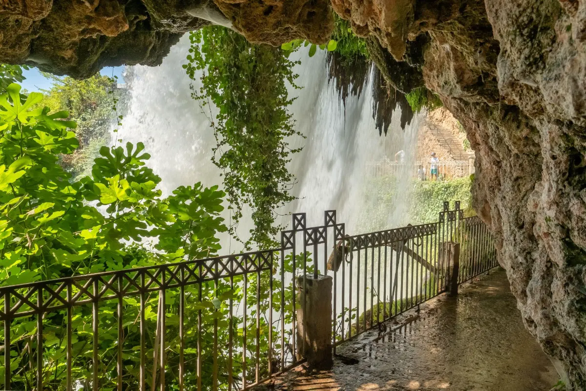 Walkway behind a cascading waterfall, framed by hanging vines and rock, with visitors viewing from a platform beyond.