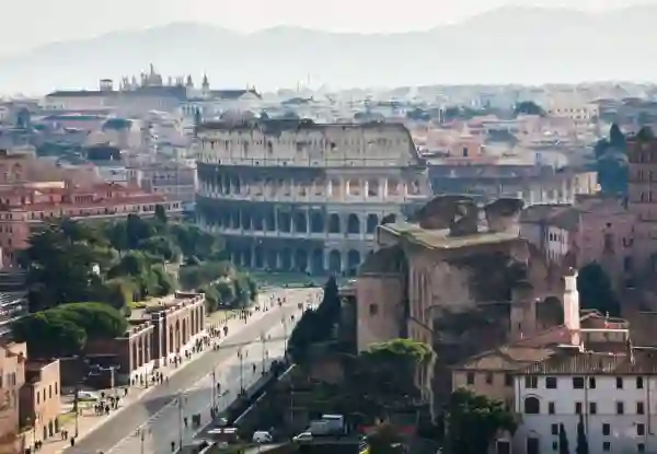 Aerial view of Rome's Colosseum rising above the historic Via dei Fori Imperiali, with pine trees and pedestrians lining the ancient boulevard. Weather In Rome In December 1